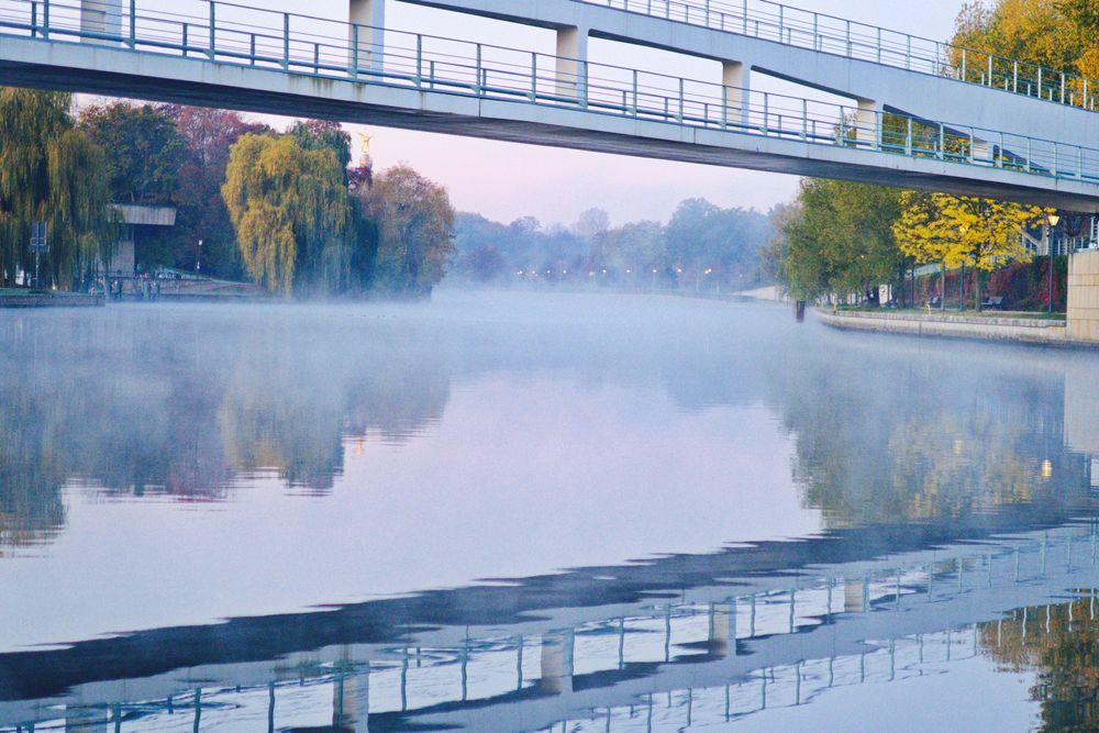 Spreebrücke am Kanzleramt morgens bei glatter Spree, so dass sich die Brücke im Wasser spiegelt. Ein frostiger Morgen, es hängt Nebel über dem Wasser.