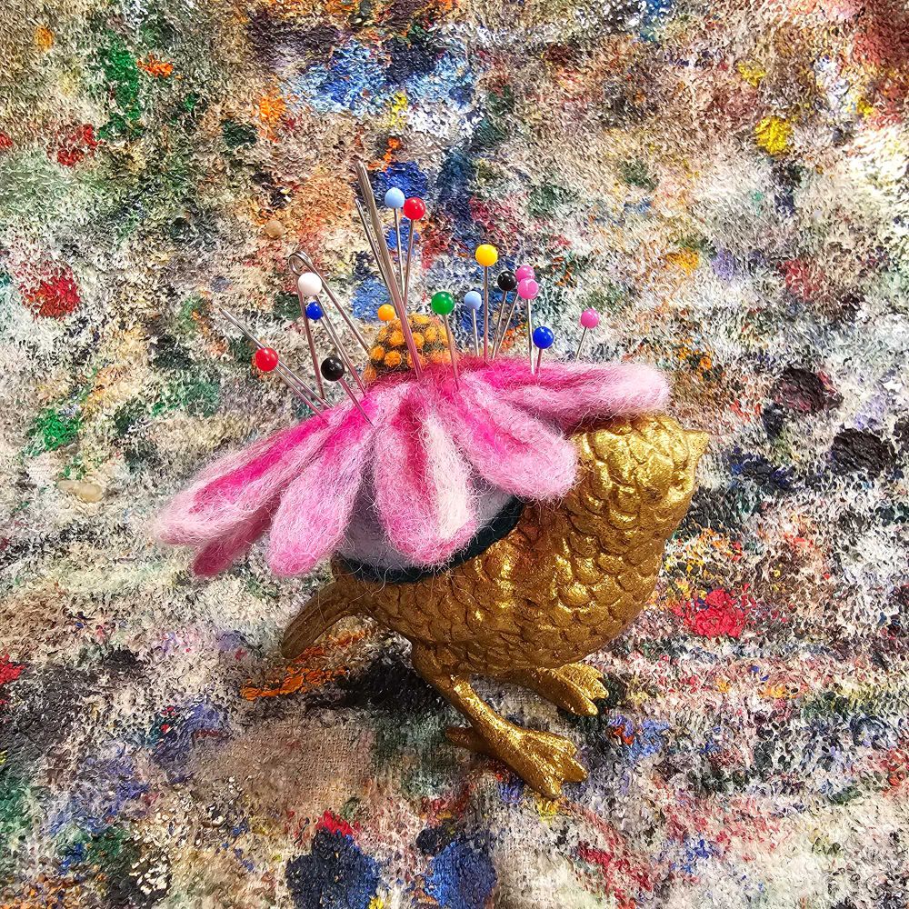 Back view: A metal song bird painted to look like brass with a purple conflower for it's back. The purple cone flower is made of wool and  functions as a pin cushion. The pincushion has multiple different straight pins and sewing needles stuck in it.  The backdrop is a Terry cloth rag that is covered on random paint.