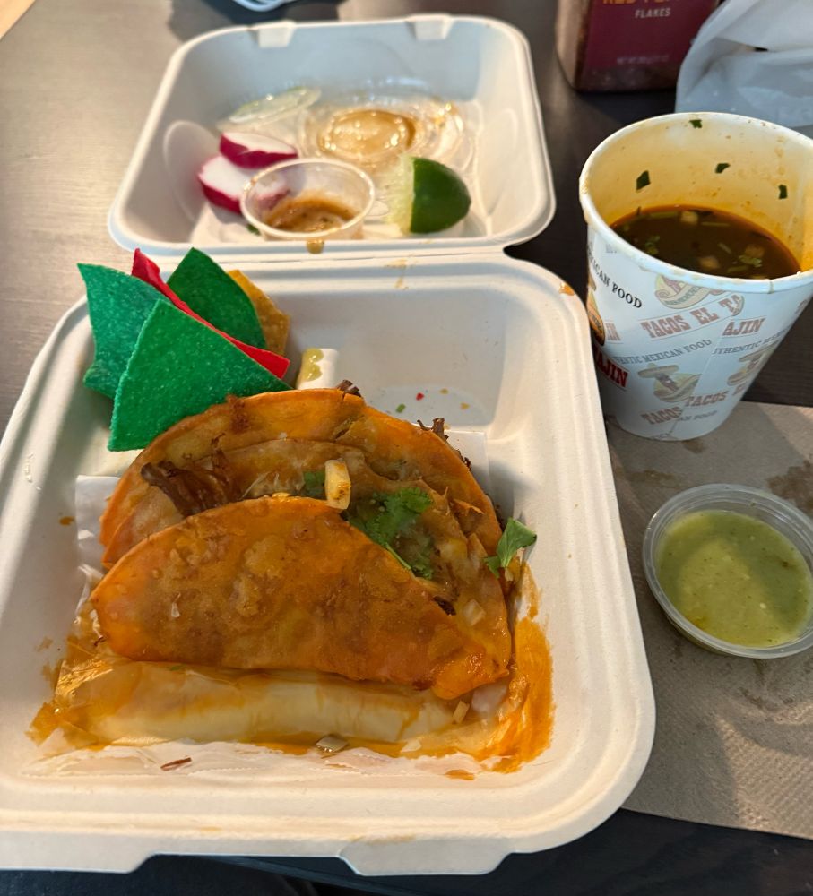 Cheese birria tacos in a to-go box. There are multicolored chips in one of the side areas. In the lid, discarded radishes, a sauce container, and a lime. To the right, the consommé (the birria) and a container of green sauce.