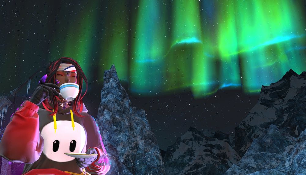 Taliya with a black and red Fall Guys hoodie is sipping a warm drink while the Coerthas aurora shines over the mountains behind her.