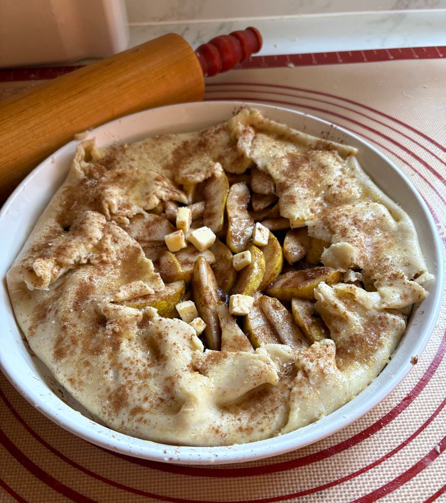 A pear galette in a white ceramic baking dish with edges folded up towards center & smothered in cinnamon ready to go into oven. A vintage rolling pin with red wooden handles peaks out from behind (it was my moms & is very beloved & used frequently)