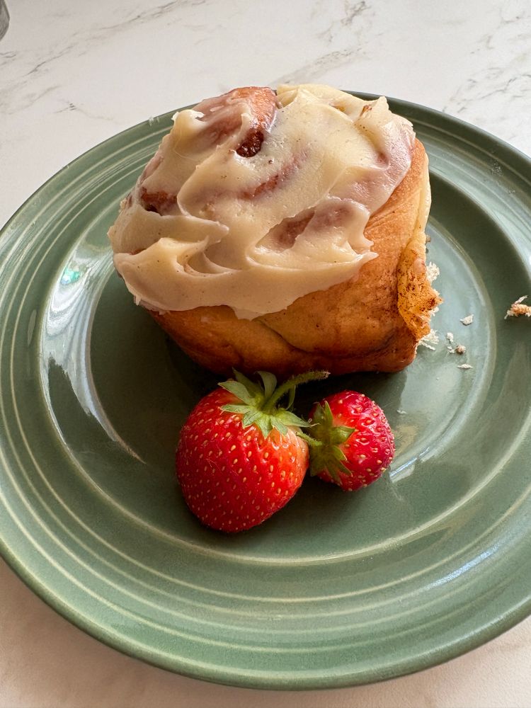 A green ceramic dish with a cinnamon rolls covered with white cream cheese frosting, Two ripe red strawberries sit on the side of the plate. Any day that starts with a fresh homemade cinnamon roll is a food day! 