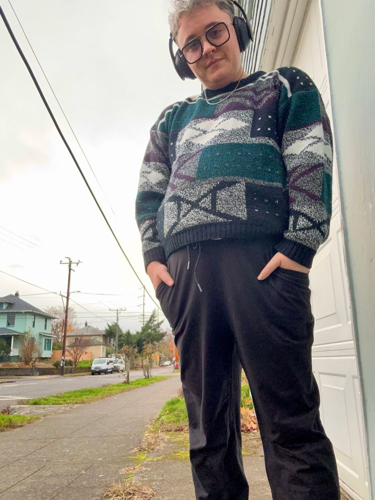 A full body shot of Ryan in the same outfit. This time you can see his black joggers as well and more of the design on the sweater. It is a black, grey, dark green and dark purple pattern. His hands are casually in the pockets of his pants. He has the same warm smile