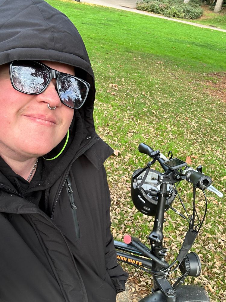 Ryan (a white trans man) stands in a black parka with the hood up next to a black e-bike with a helmet draped on it. He is wearing black sunglasses and smiling. There is green grass covered with fallen brown leaves behind him. 