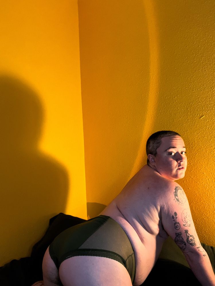 White trans man wearing dark green panties on a dark green bed spread. 