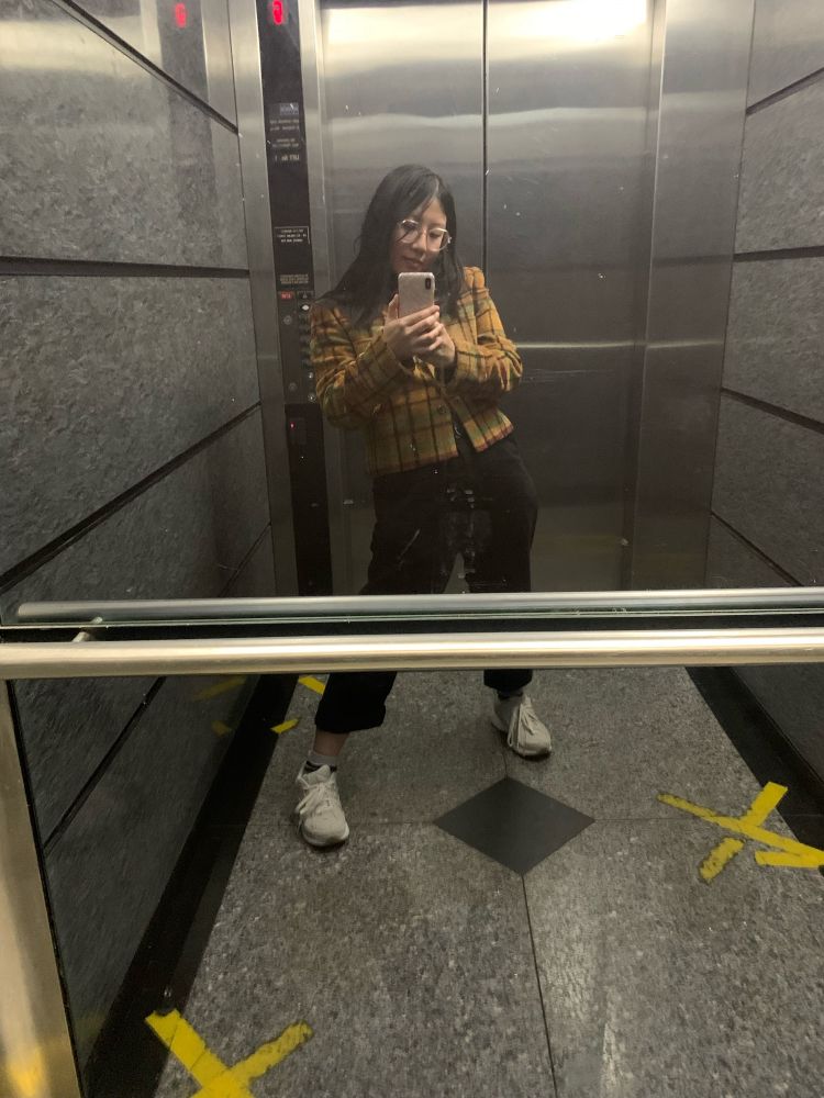 Asian person in brown check tweed jacket, baggy black pants and white sneakers in a full body mirror selfie inside a lift. They have shoulder length black hair and glasses