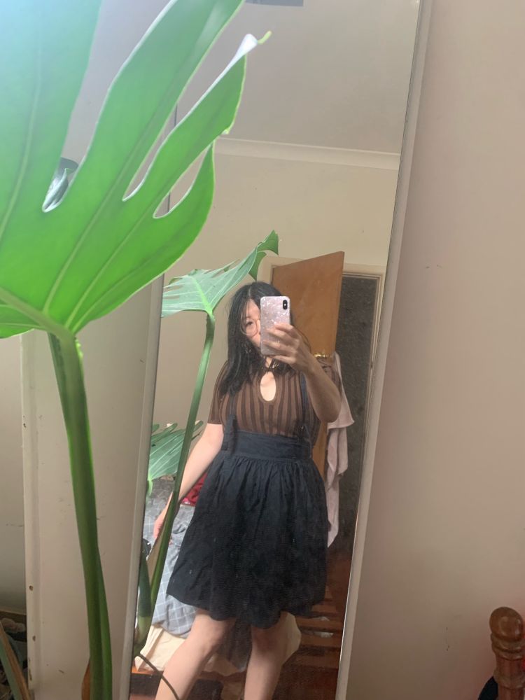 Non binary person in knit brown top and black flared skirt with straps takes selfie in a mirror