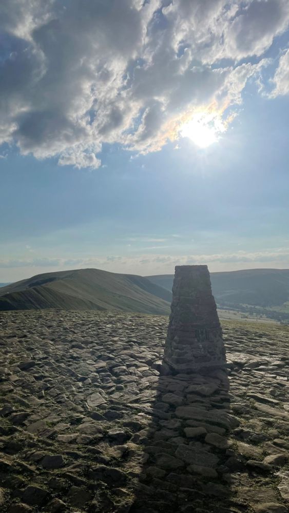 A photograph of a Trig Point in the Peak District with the late evening sun visible below the clouds