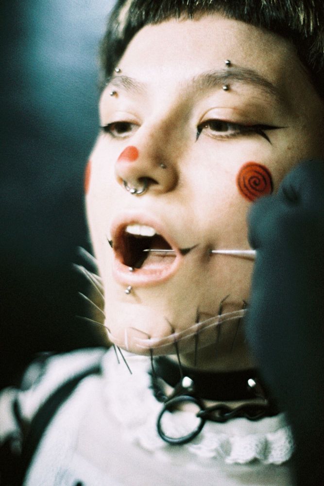 A very large needle punctures through a person's cheek. Their mouth is open, revealing the needle inside their mouth.