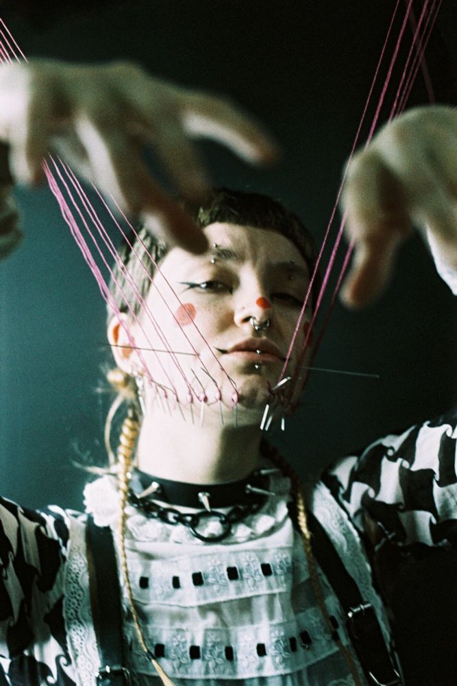 A person looks at the camera, their cheeks and hands suspended in the air by thread tied to needles protruding from their flesh.