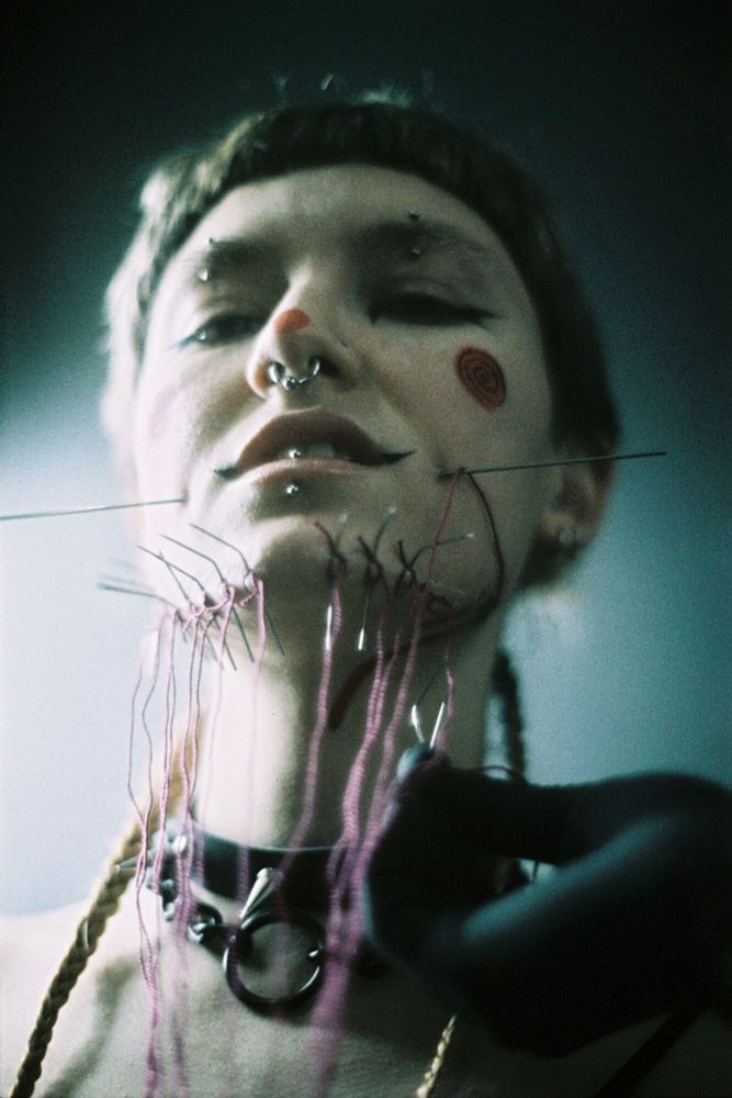 A person smirks at the camera, their cheeks skewered by a large needle and several smaller needles protruding from their jawline.
