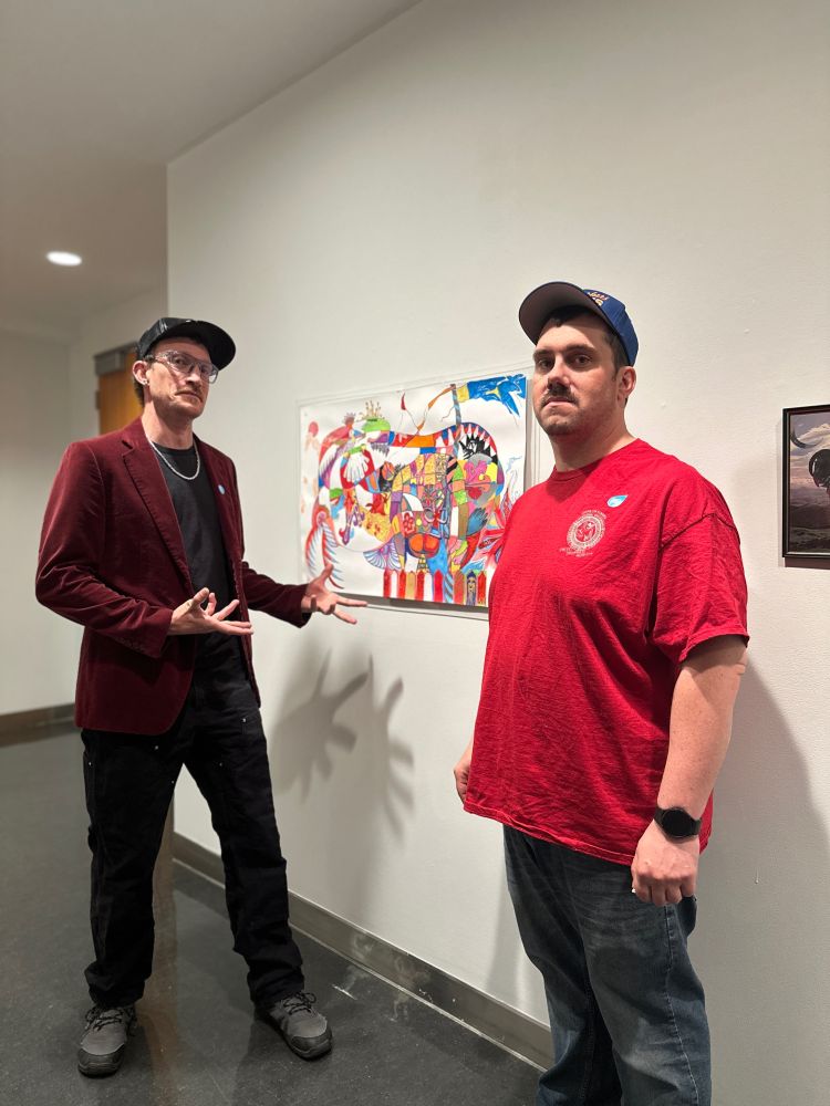 A photo of Finn posing in a velvet jacket beside his painting Beastro Praha, beside a friend. Both are wearing baseball caps. 