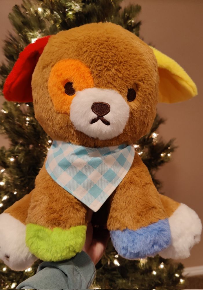 a picture of a large rainbow dog plush wearing a blue bandana. in the background there is a christmas tree.