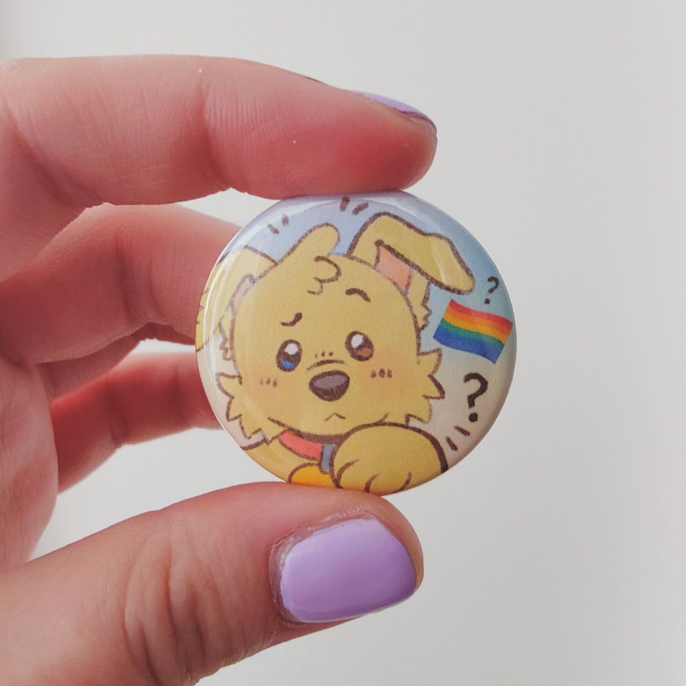 a photo of a tinplate button badge featuring artwork of a puppy looking confused, with a pride flag emoji and some question marks