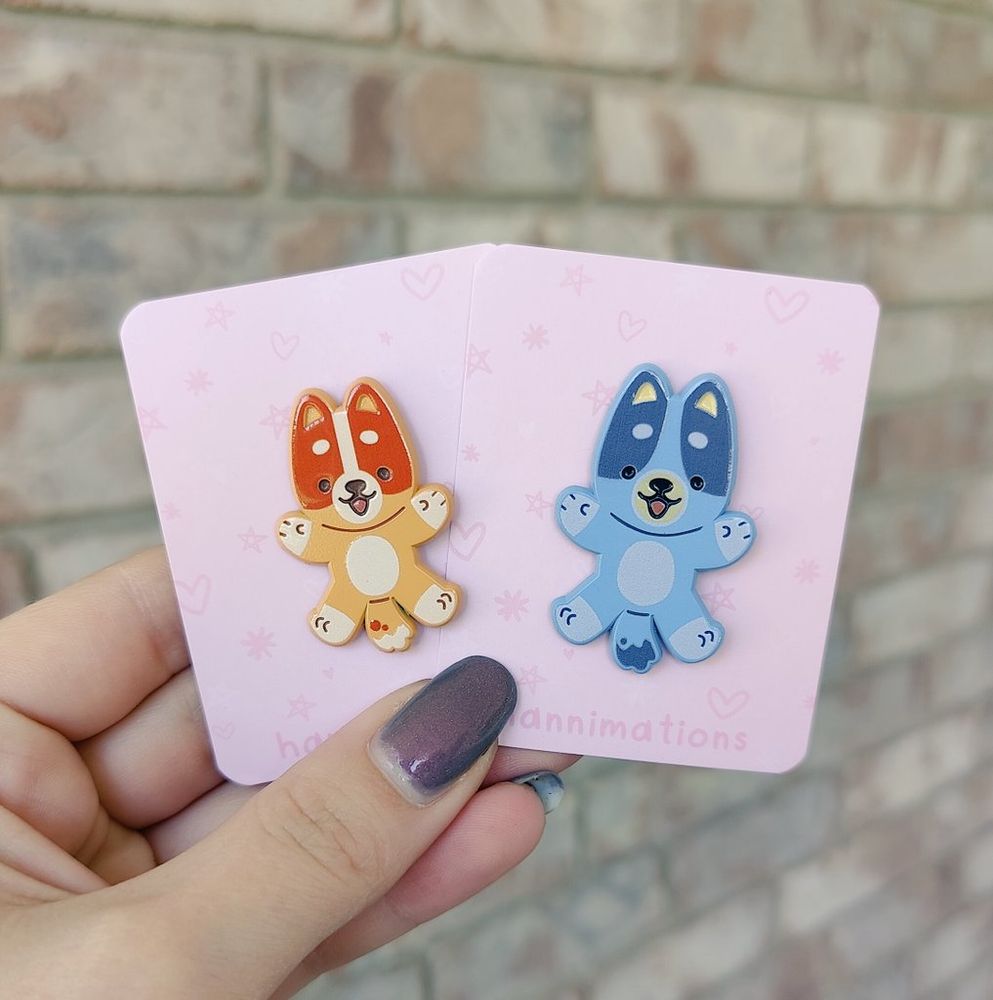 a photo of a hand holding two enamel pins, each on a pink backing paper. the pin on the left resembles bingo from the TV show bluey, and the pin on the right resembles bluey from the same show.
