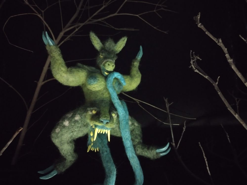 a felted puppet with bizarre anatomy. it is humanoid with a donkey head, it has hooves for feet, and hooves for hands, and an oversized, long-toothed, cat-like face growing out of its crotch. Instead of using its legs, it stands on the cat's tongue. the tongue is busy with the creature's own breast.