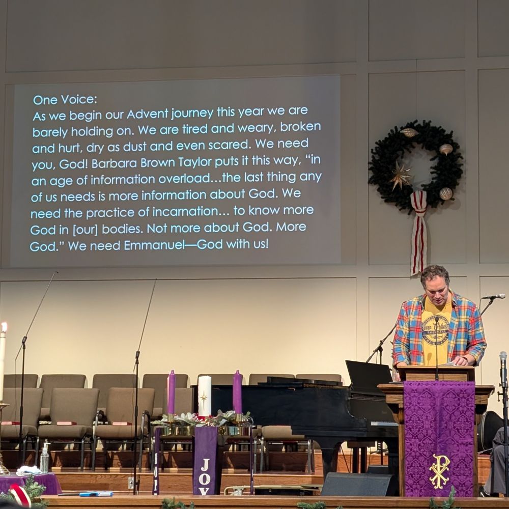 A casually dressed preacher at a lectern. Advent wreath in foreground. Projected on the back wall is a responsive reading including a quote from Barbara Brown Taylor" in an age of information overload. The last thing any of us needs is more information about God. We need the practice of incarnation to know more God in our bodies not more about God, more God we need Emmanuel."