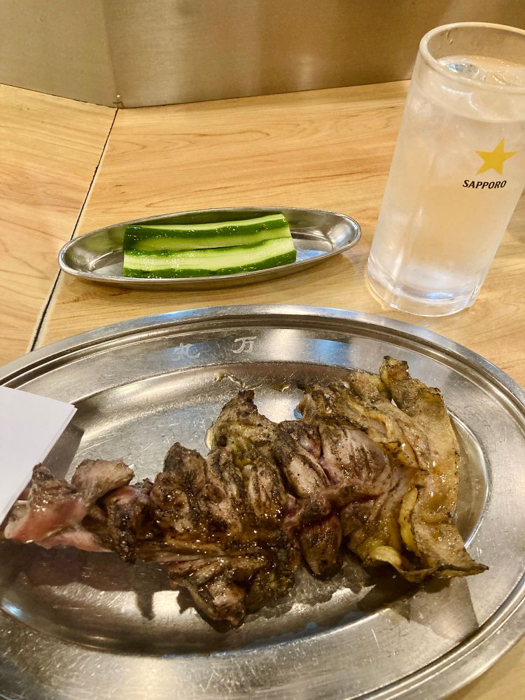 骨付き鶏のもも焼き、骨の近くはほとんど赤い。付け合わせの半割きゅうりと焼酎の水割り。