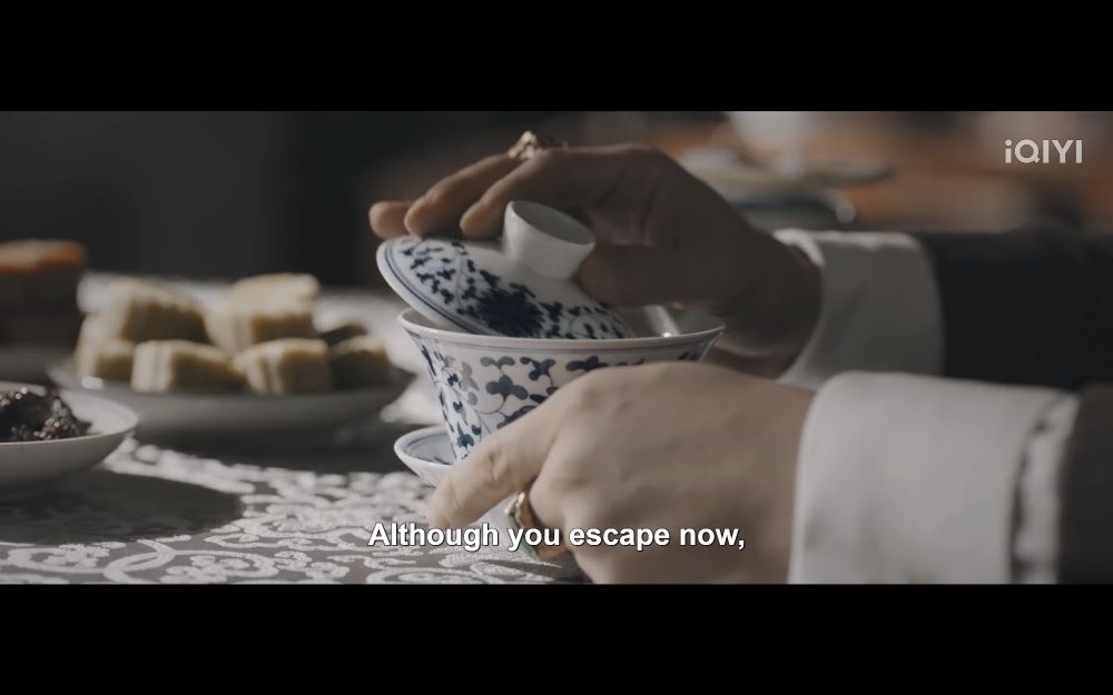 Cheng Feng Tai holds a teacup on a table full of pastries, skimming the top of the tea's surface with the lid of the cup