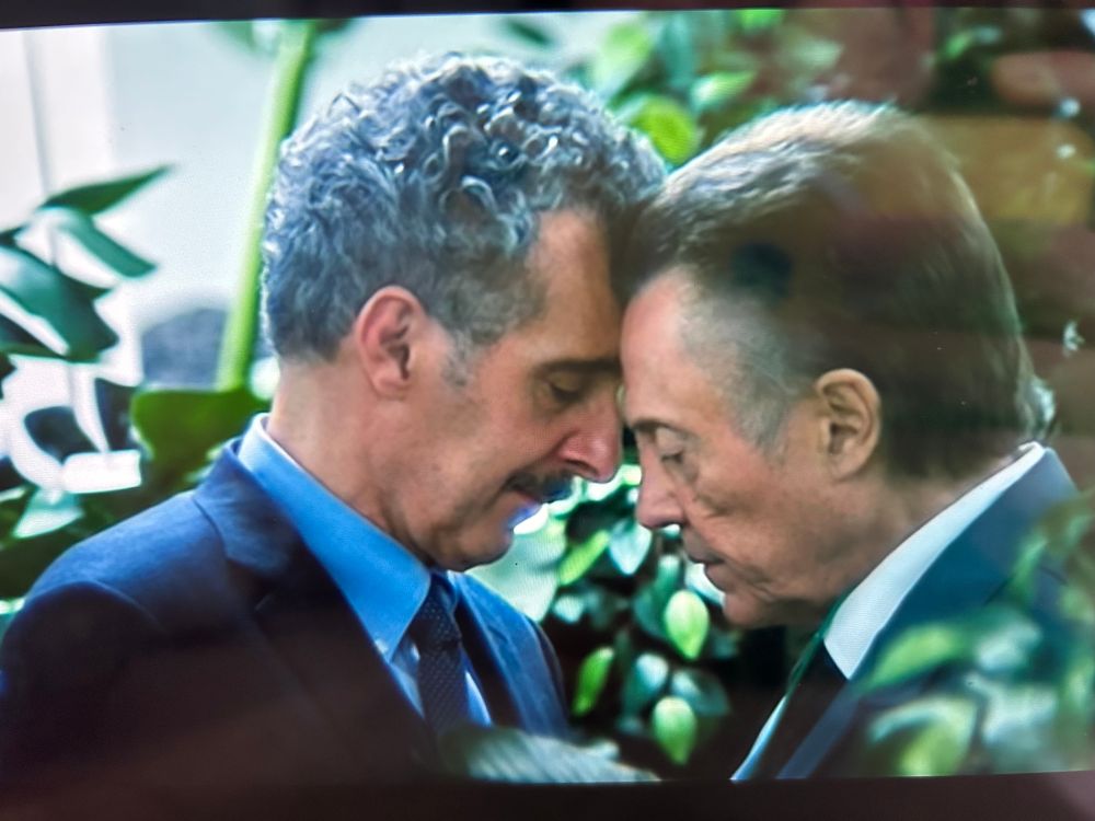 Two older white men, one with short and curly grey hair, the other short and straight dark grey hair, press their foreheads together with their eyes closed. They are surrounded my various luscious green plants. They are in love.
