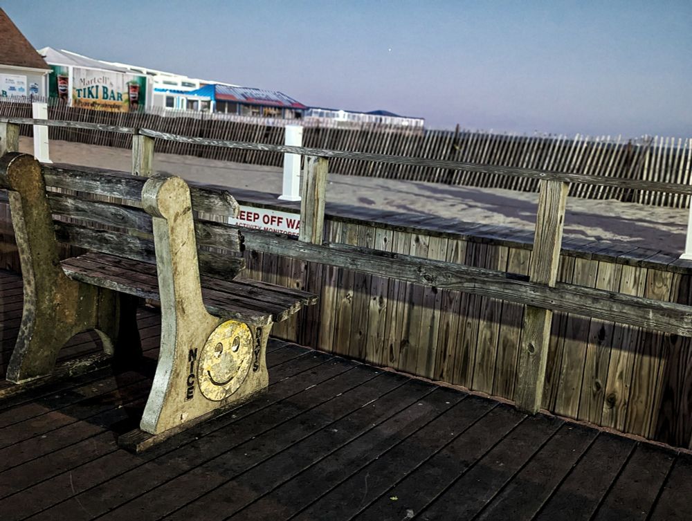 A bench on a boardwalk, the side of the bench has a painting of a smiley face and the words "Nice Days." There is a sand fence in front of the bench and a row of tents in the background that are closed up for the winter. 