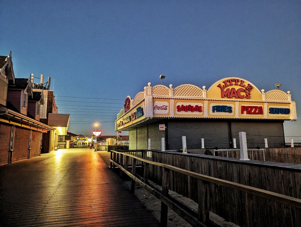 An empty boardwalk with shuttered buildings, the main one in focus is "Little Mac's" featuring vintage looking words describing what they sell, "Coca Cola, Sausage, Fries, Pizza and Seafood."