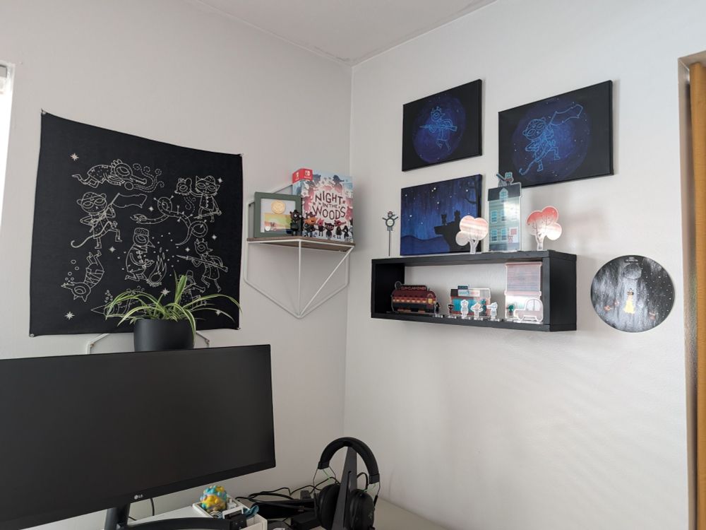 The shelf and paintings described in the other photograph of this set, as well as two other small shelves in front of a computer monitor. One shelf contains a spider plant behind a printed bandana showing the constellations from the game Night in the Woods. The other shelf is displaying the box of the Collector's edition of the same game, a coin and card from that box, and individual figurines of Angus and Gregg.