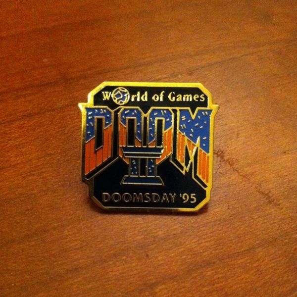 pin badge with 'world of games' in gold at the top in small text (and the 'o' looks like the earth). below this is the doom 2 font, top half of the letters is dark blue, bottom half orange and the II below is in dark blue. below this, at the bottom of the badge is the bolt gold text 'doomsday '95'.