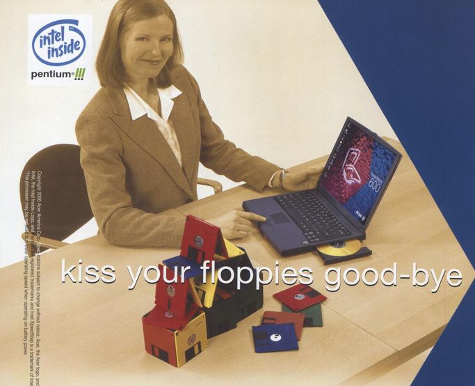 intel inside pentium III logo in the upper left hand corner of the image.
a woman wearing a business suit is using a laptop with a cd-rw ejected (she is smiling and looking up at the camera). floppy disk are laying on the desk and are also stacked up in a structure that could very easily be toppled over.
text: kiss your floppies good-bye