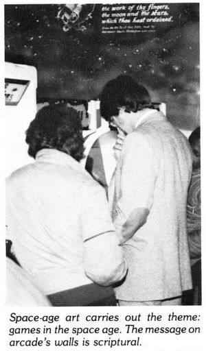 two adults with their backs turned to an arcade machine. the wall looks like space and stars. there is some writing printed on it: ...the work of thy fingers, the moon and the strs which then hest(?) ordained. (something too small and indecipherable underneath).