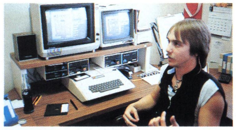 richard garriott, straight brown hair, mustache, black vest, sitting at a workstation, 2 screens on a raised platform, a keyboard computer and disk drives below, a floppy disk on the desk.