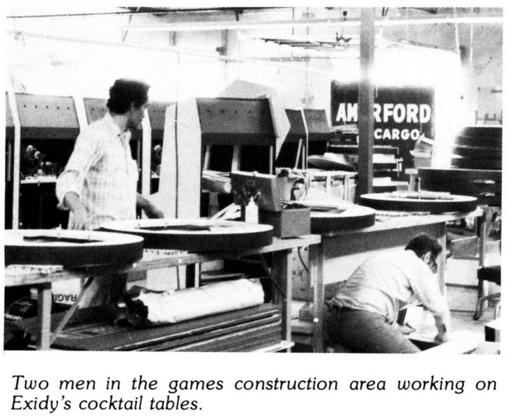 black and white photo of two men working on the construction of arcade cocktail tables (the top round parts with cut outs inside can be seen in a line on benches). stand up arcade cabinets can be in the background.
caption: two men in the games construction area working on exidy's cocktail tables.