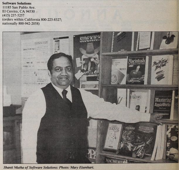 a man named shanti mutha stands in front of a display of books, some macintosh books and old game hint books (or software boxes?) such as infocom's deadline and seastalker.