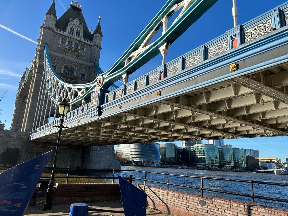 Showing underside of Tower Bridge, square girders, blue and gold railings, one tower and a very blue sky