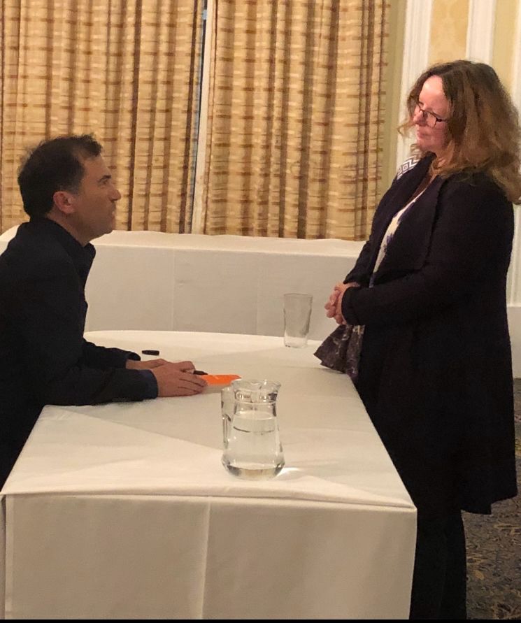 Ros Atkins sitting at a table, signing his book, with me looking and listening