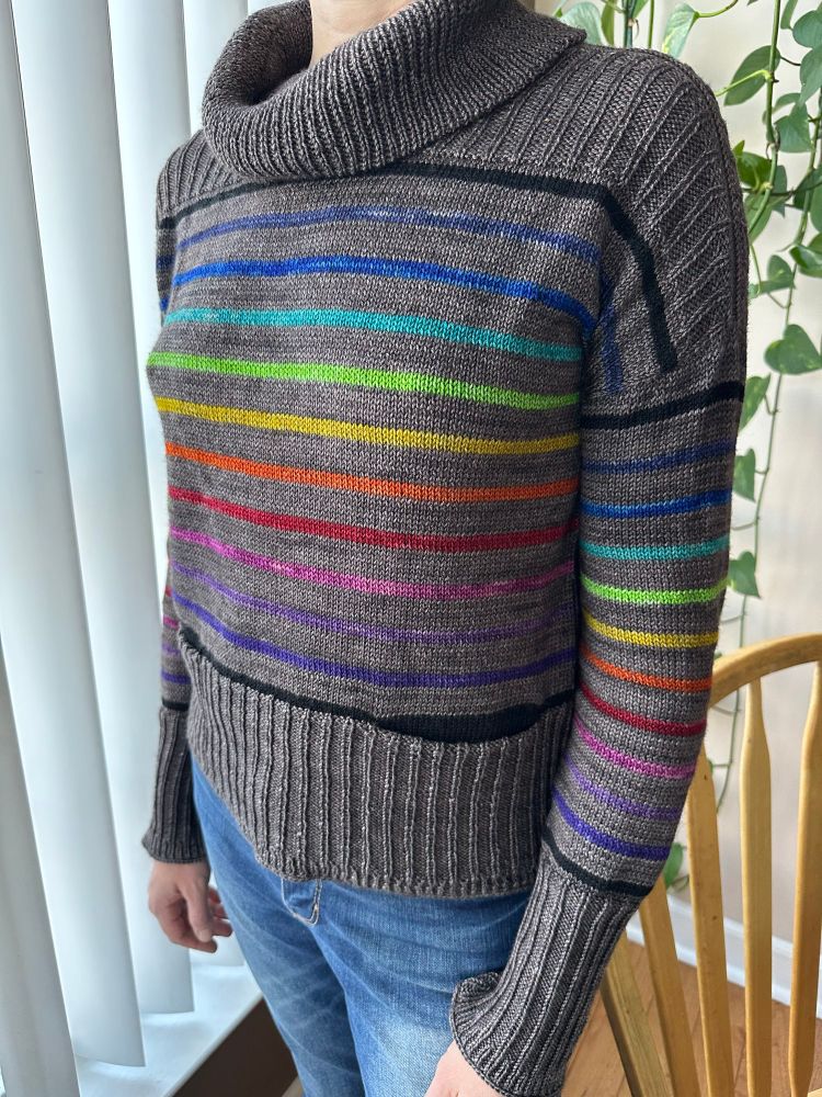 Slightly side facing view of a dark grey sweater with rainbow stripes and wide ribbed edges with a cowl neck and pockets.