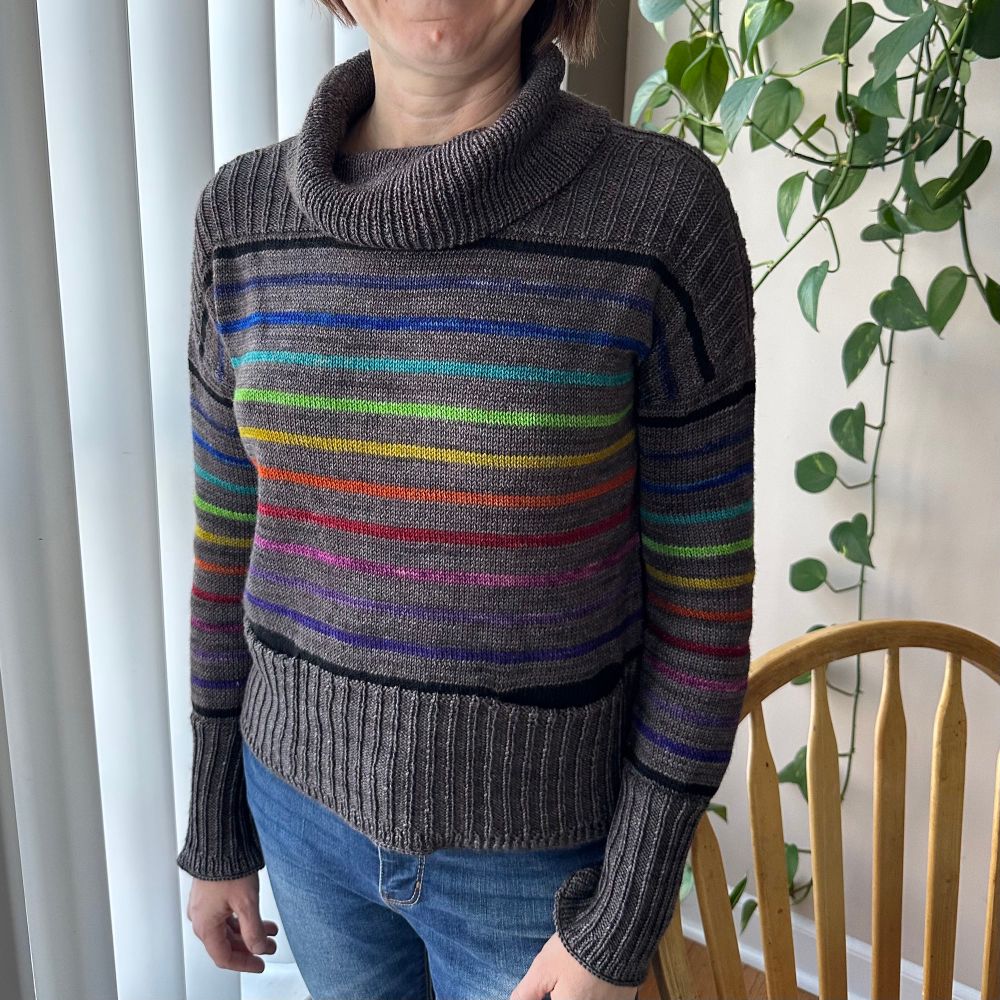 Front facing view of a dark grey sweater with rainbow stripes and wide ribbed edges with a cowl neck and pockets.