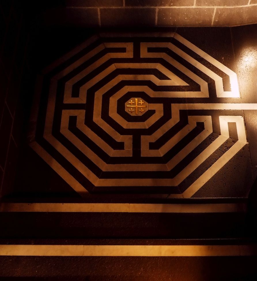 Eine Treppe aus dunklem Stein biegt auf einem quadratischen Absatz rechtwinklig ab. Auf diesem Absatz befindet sich in hellen Linien ein achteckiges Bodenlabyrinth mit einem kleinen goldenen Jerusalemkreuz im Zentrum. Eine helle Linie führt vom rechten Bildrand in das Labyrinth hinein. Auch die Stirnseiten der Treppenstufen sind oben mit hellen Streifen abgesetzt, um im Halbdunkel beim Gehen nicht zu stolpern.
