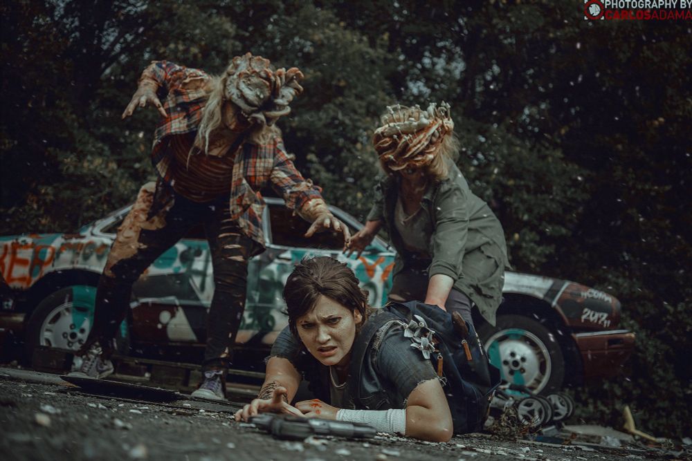 Abi is cosplaying Ellie Williams. She is crawlig towards a gun, two clickers (Hattie and Clara) are pursuing her.