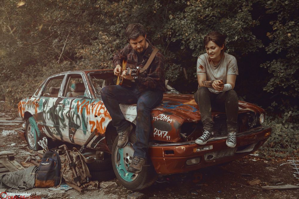 Ben and Abi are cosplaying Joel and Ellie. They are sat on the bonnet of a burnt out car. Joel is playing guitar and Ellie is singing.