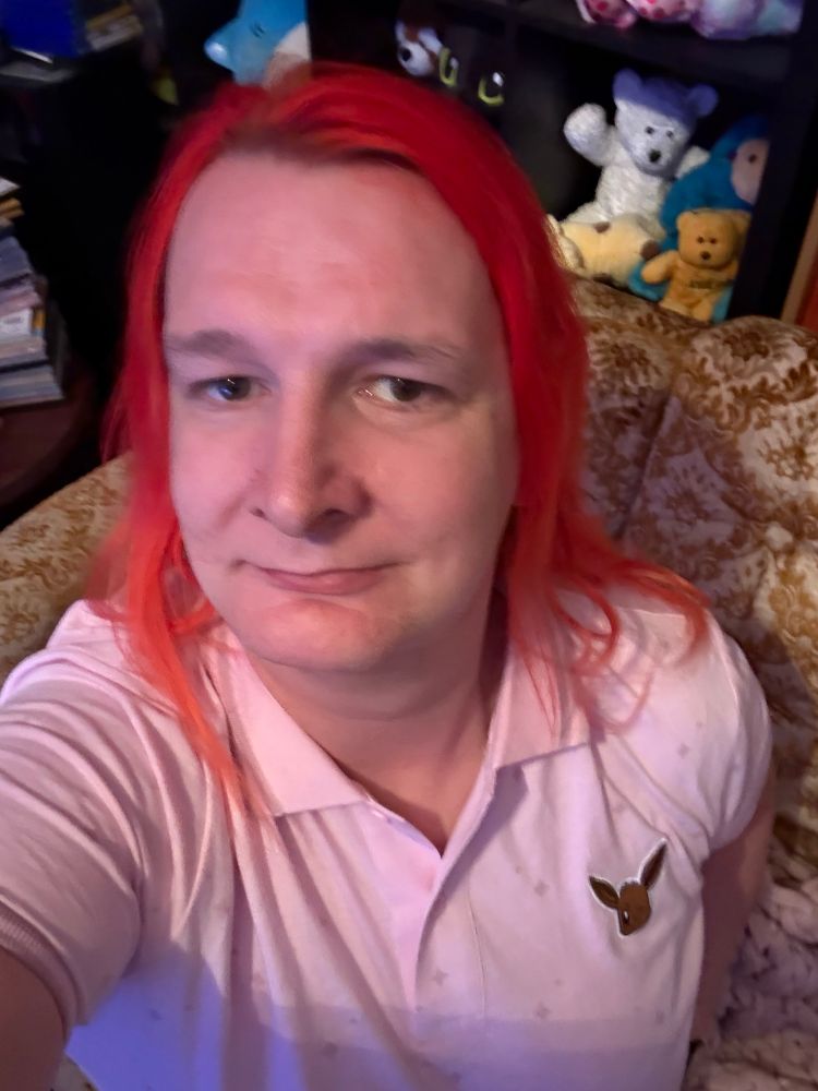 Selfie of myself in a pink work shirt with an image of an Eevee head on it. From the video game pokemon. 