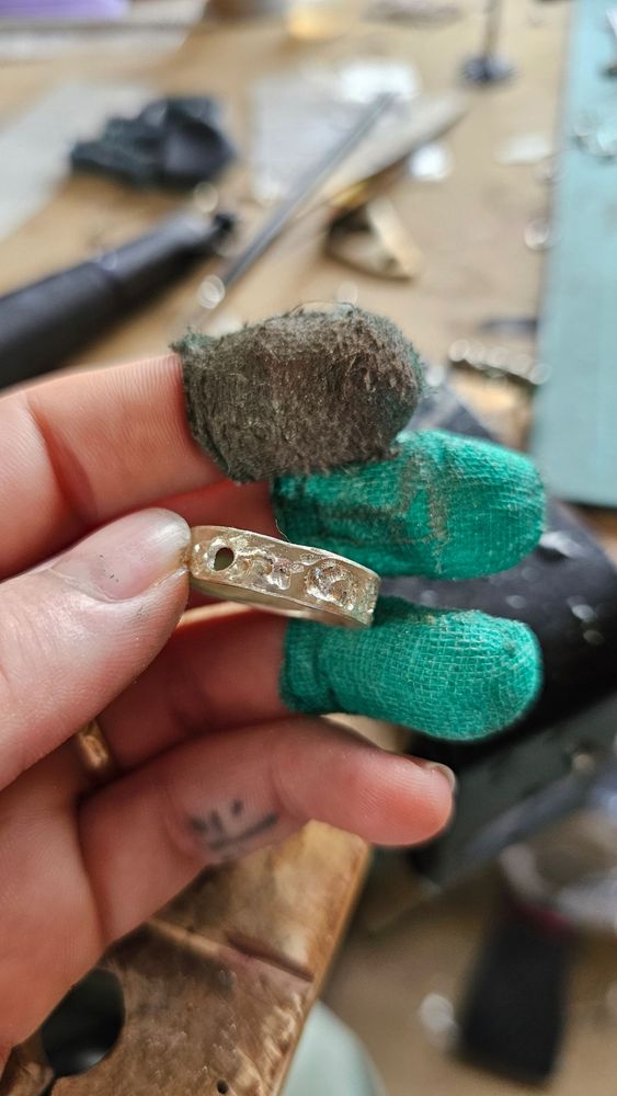 A hand with green protective tape on the fingers holds a silver ring in the process of being carved. The carved pits and lines are a brighter white than the rest of the large silver band.