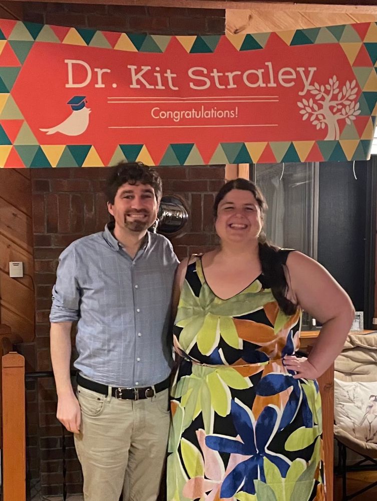 Two people standing under a colorful banner that reads “Dr. Kit Straley congratulations!” The banner has a bird with a graduation style cap.