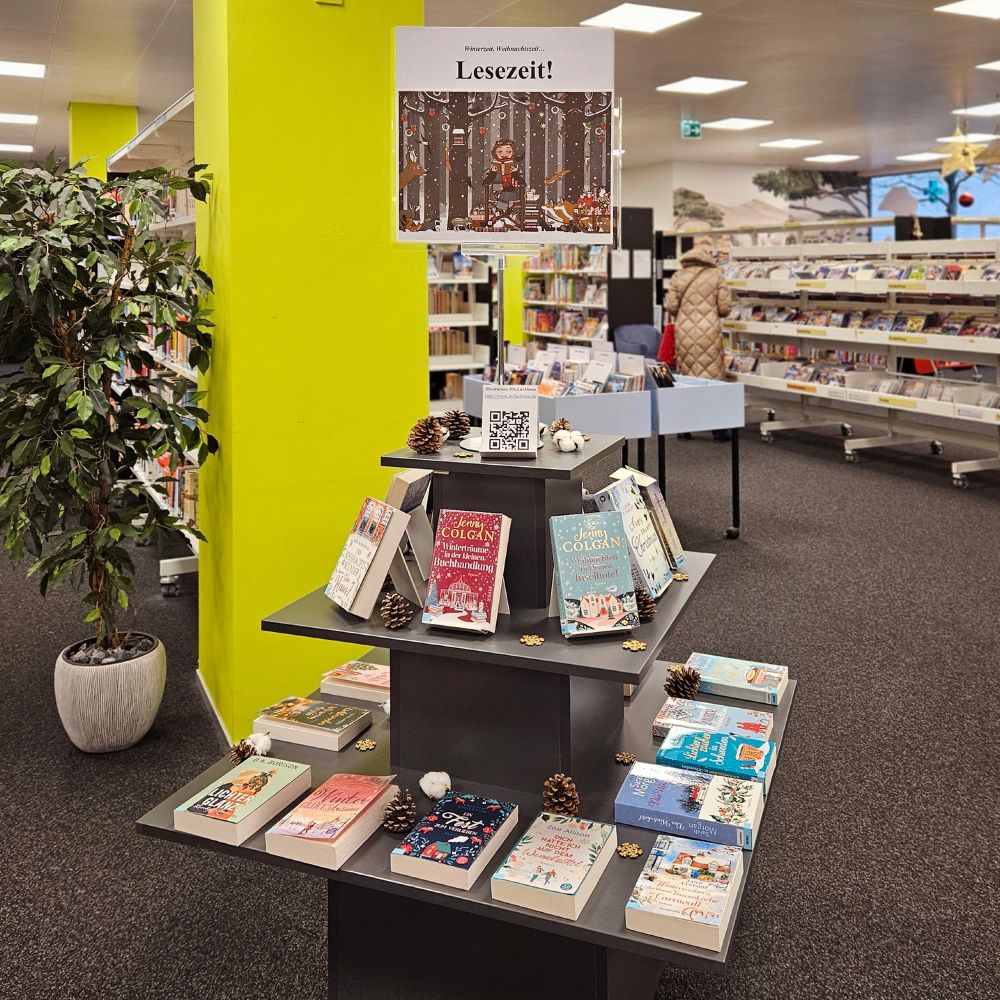 Foto des Etagen-Tisches in der Stadt- und Regionalbibliothek Uster, auf dem weihnachtliche Romane präsentiert werden – gekrönt von einem Poster mit einer Illustration der von Iris Luckhaus gezeichneten Figur Lily Lux, die gemütlich im festlich geschmückten Winterwald sitzt und den Tieren vorliest. 