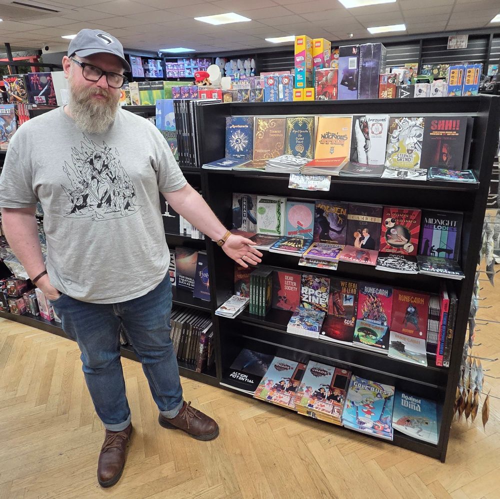 Me (Colin), a white man with glasses and a beard, points at a shelf full of By Odin's Beard RPG books, amidst a bunch of other great indie titles.