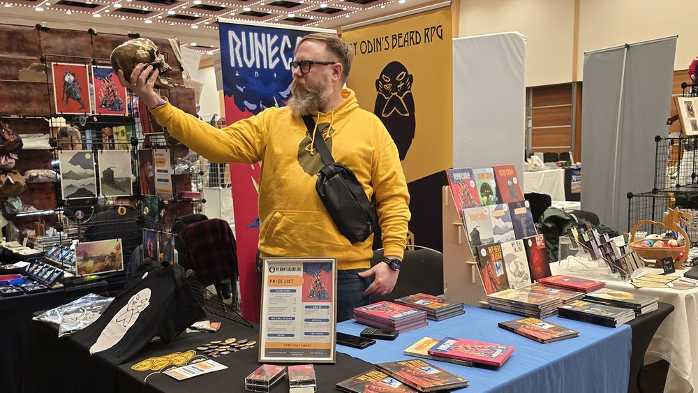 Me (Colin), a white man with beard and glasses, holds a bronze skull like Hamlet behind the By Odin's Beard RPG table at Dragonmeet 2024.