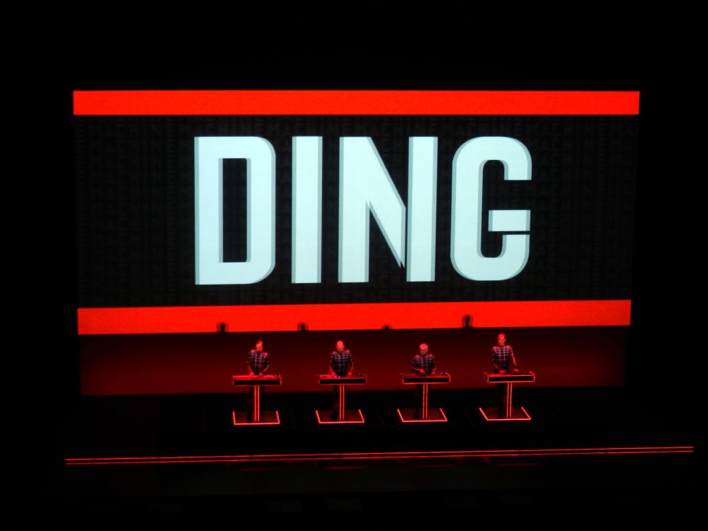 Dieses Bild zeigt eine Konzertbühne während einer Live-Performance der Band Kraftwerk.

    Bühnendesign: Im Hintergrund dominiert eine große, rechteckige Videowand.

        Die Wand ist von einem auffälligen roten Rahmen umgeben.

        In der Mitte wird in großen, blockartigen, hellgrau-weißen Buchstaben das Wort "DING" angezeigt.

     Performer: Auf der Bühne stehen vier Performer nebeneinander, alle ähnlich gekleidet in karierten Langarmhemden.

        Jeder Performer steht hinter einem separaten, pultartigen Gerät (vermutlich Synthesizer oder Controller).

        Die Pults haben eine klare, metallische, rot beleuchtete Struktur.

    Beleuchtung: Die Beleuchtung ist dramatisch und minimalistisch, dominiert von Rot- und Schwarztönen, was die futuristisch-technologische Atmosphäre der Performance unterstreicht.

