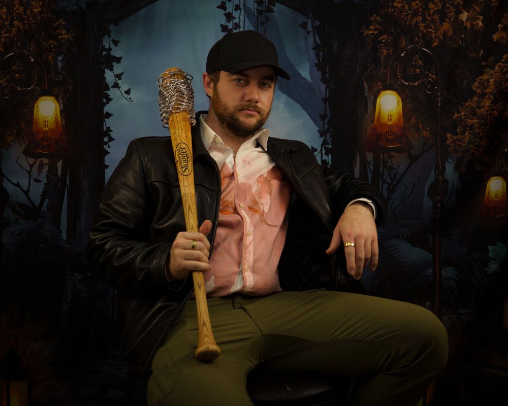 Zach sitting in a chair wearing a blood-stained white button up shirt, black leather jacket, green pants, and a black hat. He is holding a bloody wooden baseball bat with chain wrapped on the end 