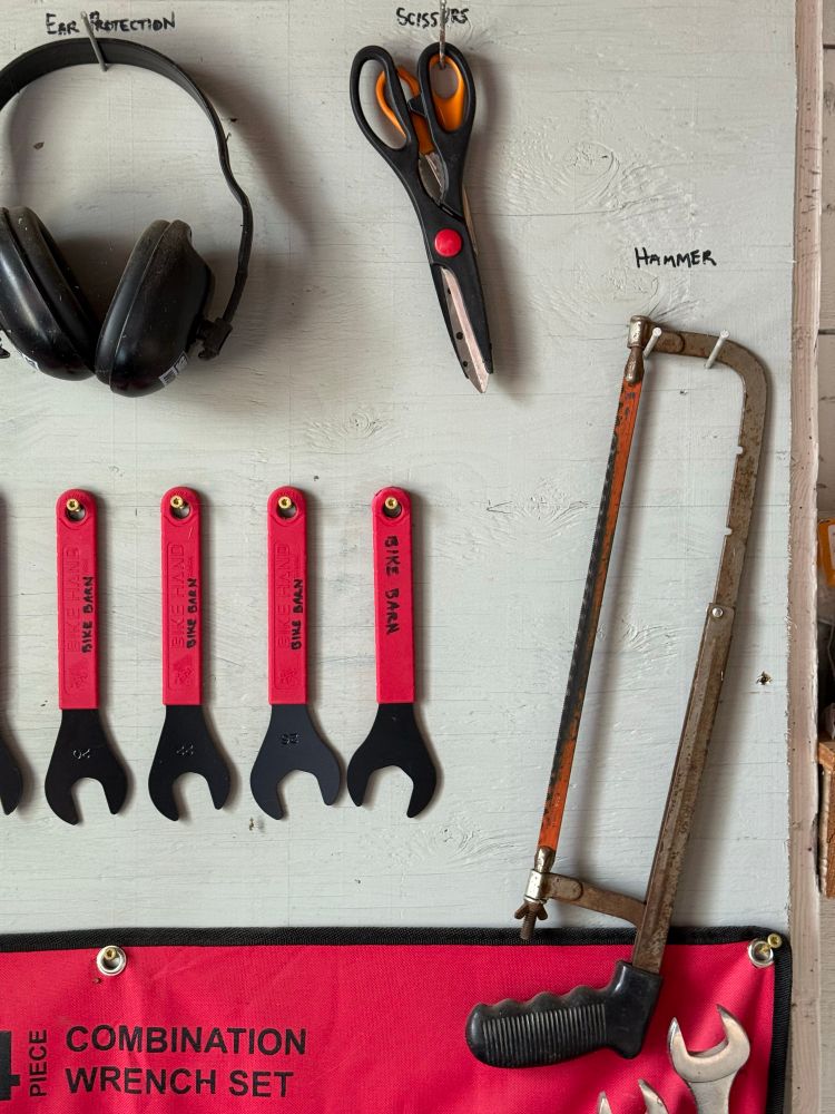 Tools hanging on a wall, labeled wrenches, scissors, ear protection, and hammer. But instead of a hammer it’s a saw.