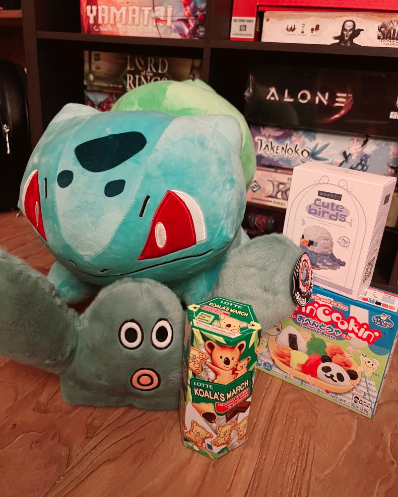 The photo is of a few arcade prizes, including a large bulbasaur plush, a small dark green monster plush, koala cookies, a poppin' cookin' set, and a bird block set.
